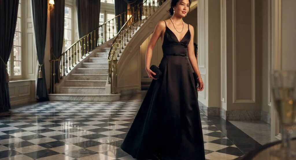 Woman in a black satin black-tie evening gown with pearl necklace, standing near a grand staircase